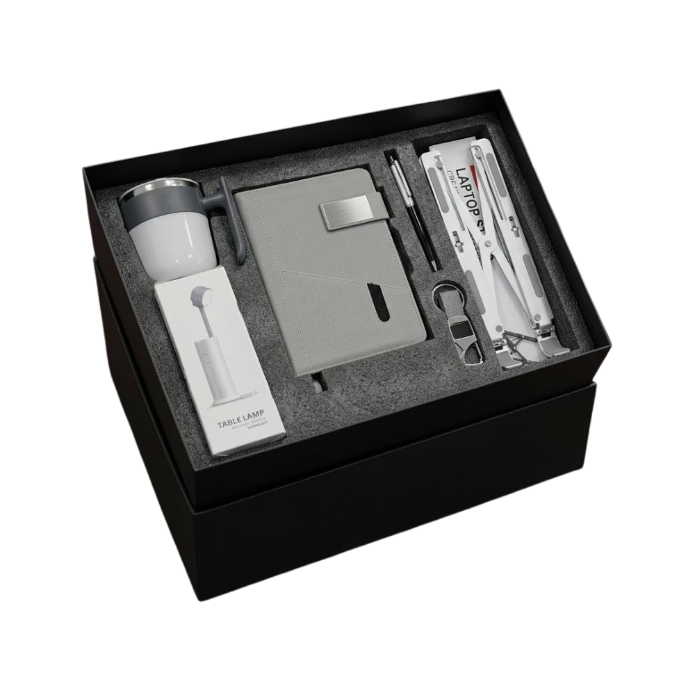 A premium black gift box containing a grey notebook, mug, pen, keychain, laptop stand, and a table lamp.